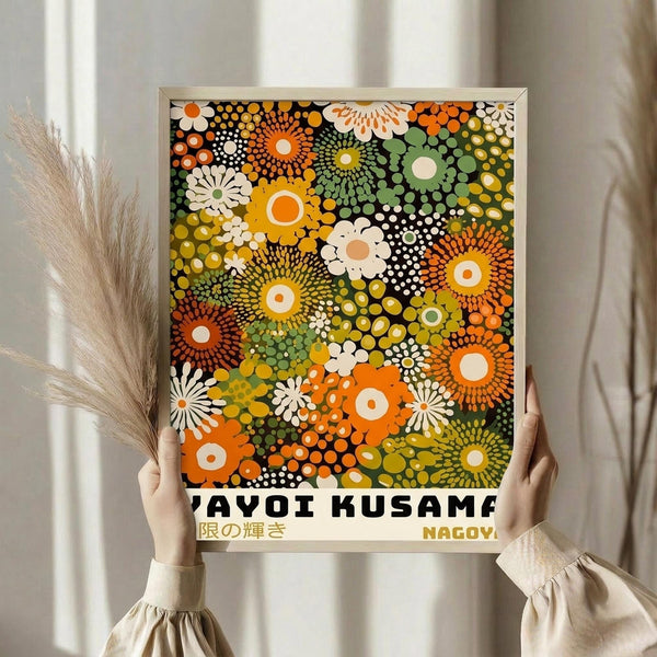 May include: Person holding framed art print featuring colorful dotted floral pattern in orange, green, yellow, and white circles against dark background. Japanese text visible at bottom. Natural lighting with dried wheat stalks and neutral curtains in background. Hands wearing cream colored ruffled sleeves grip wooden frame edges.