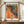 May include: Hands holding framed poster featuring vibrant abstract artwork with swirling orange and yellow organic forms on left side flowing into colorful polka dot pattern on right against black background. Bold black text reads "YAYOI KUSAMA" at top and "TOKYO" at bottom. Cream colored frame sits on wooden surface next to white ceramic vase and stacked books against white paneled wall.