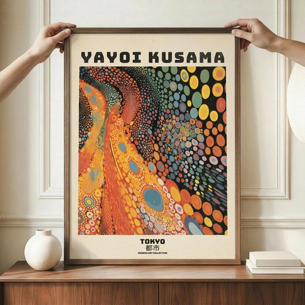 May include: Hands holding framed poster featuring vibrant abstract artwork with swirling orange and yellow organic forms on left side flowing into colorful polka dot pattern on right against black background. Bold black text reads "YAYOI KUSAMA" at top and "TOKYO" at bottom. Cream colored frame sits on wooden surface next to white ceramic vase and stacked books against white paneled wall.