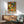 May include: Framed poster with colorful abstract pattern of overlapping circles, dots, and flower-like shapes in orange, green, yellow, and white tones, displayed above a wooden sideboard with white ceramic lamp, next to a sage green mid-century modern armchair with wooden arms in a bright, minimalist room with white walls and natural lighting.