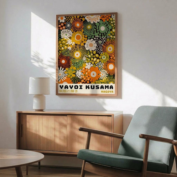 May include: Framed poster with colorful abstract pattern of overlapping circles, dots, and flower-like shapes in orange, green, yellow, and white tones, displayed above a wooden sideboard with white ceramic lamp, next to a sage green mid-century modern armchair with wooden arms in a bright, minimalist room with white walls and natural lighting.