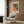 May include: Colorful abstract art poster with orange, green, and multicolored dotted pattern hanging on wood-paneled wall above beige upholstered armchair with geometric pillow. Gold floor lamp and dark side table with open book nearby. Modern minimalist interior with warm wood tones and natural lighting from window with white curtains.