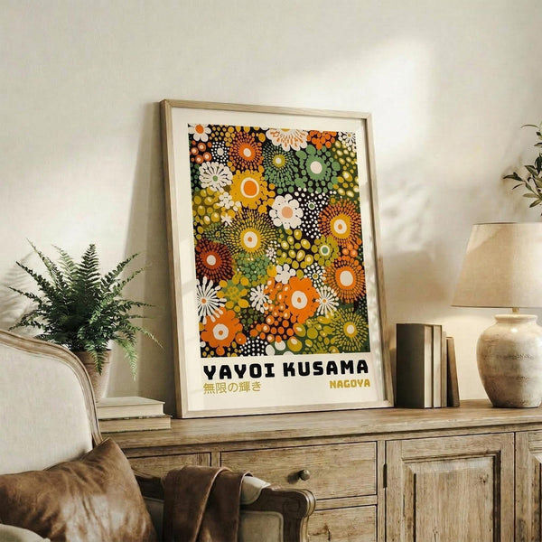 May include: Framed art print featuring colorful circular and floral patterns in orange, green, yellow, and white dots displayed on a wooden sideboard next to a fern plant, ceramic vase, and books in a neutral-toned living room setting with beige furniture.