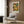 May include: Framed poster with colorful circular dot patterns in orange, green, yellow and white tones hanging on wood paneled wall above beige upholstered armchair with geometric patterned pillow, brass floor lamp, and dark round side table with open book in modern living room setting with white curtains and light wood flooring.