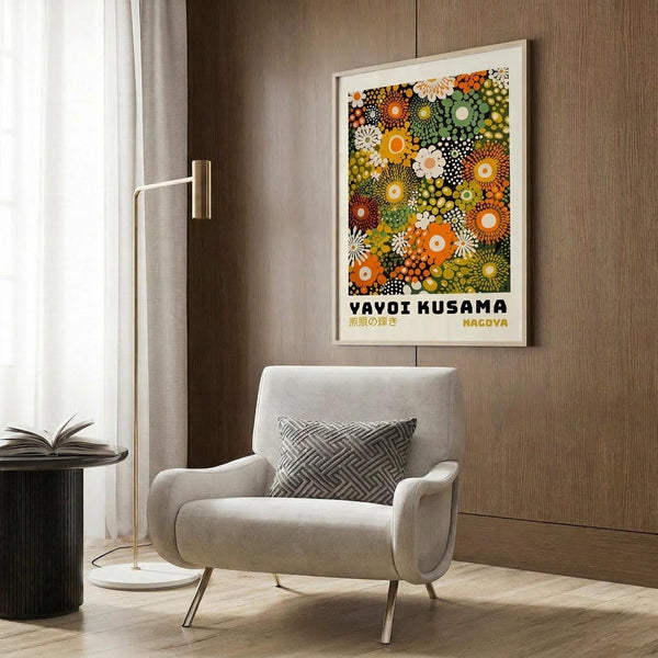 May include: Framed poster with colorful circular dot patterns in orange, green, yellow and white tones hanging on wood paneled wall above beige upholstered armchair with geometric patterned pillow, brass floor lamp, and dark round side table with open book in modern living room setting with white curtains and light wood flooring.
