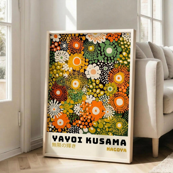 May include: Framed poster with colorful polka dot artwork in vibrant oranges, greens, yellows, and white against black background, featuring circular flower-like patterns and dots in Kusama's signature style. Text reads "YAYOI KUSAMA" with Japanese characters and "NAGOYA" below. Light wood frame leaning against white wall next to beige upholstered chair in bright room with large windows.