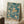 May include: Framed poster with teal blue watercolor background featuring floating jellyfish in cream and peach tones with flowing tentacles, displayed in light wooden frame leaning against white wall corner, white coffee cup on wooden block nearby, beige knit blanket visible at edge, soft natural lighting creating shadows on wall