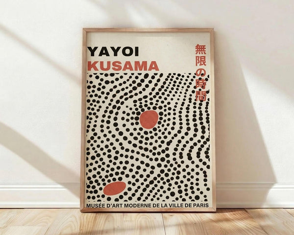 May include: Framed poster with cream background featuring black polka dot pattern radiating outward, two red oval shapes, "YAYOI KUSAMA" text at top in black and red letters, Japanese characters in red on right side, and museum text at bottom. Natural wood frame leaning against white wall on light wooden floor with soft window light shadows.