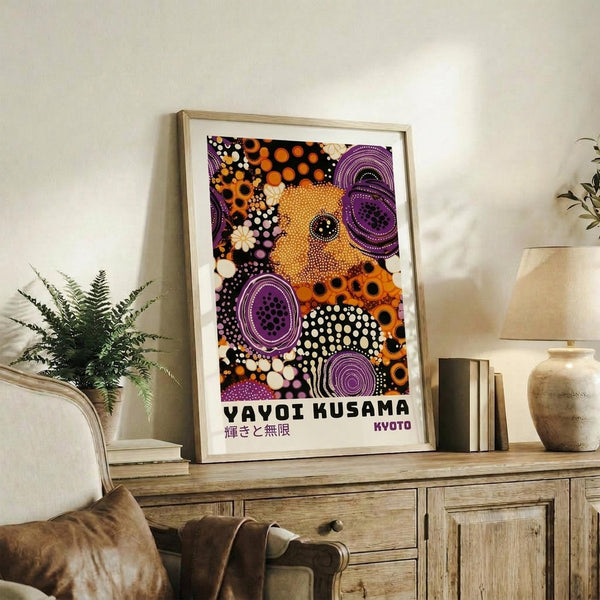 May include: Framed colorful abstract art print with orange, purple, and black polka dot patterns displayed on wooden console table beside fern plant, ceramic vase, and books in modern living room setting with beige armchair