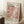 May include: Framed art poster featuring pink and coral polka dot pattern with black and red text reading "YAYOI KUSAMA" and Japanese characters, dated April 1986 Tokyo 1998. Light wood frame leaning against white wall corner on textured cream rug, with coffee cup on wooden block and beige knit throw blanket visible in cozy room setting with natural window lighting.