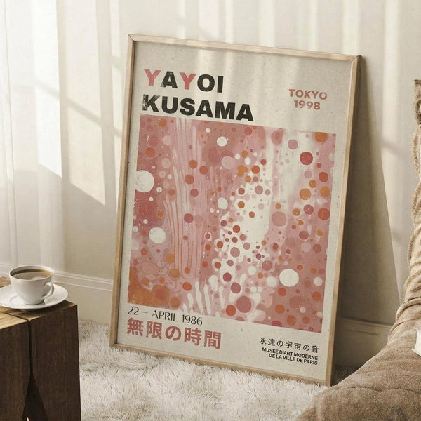 May include: Framed art poster featuring pink and coral polka dot pattern with black and red text reading "YAYOI KUSAMA" and Japanese characters, dated April 1986 Tokyo 1998. Light wood frame leaning against white wall corner on textured cream rug, with coffee cup on wooden block and beige knit throw blanket visible in cozy room setting with natural window lighting.