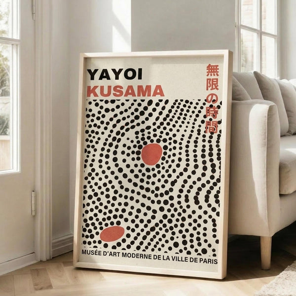 May include: Framed art print featuring "YAYOI KUSAMA" in black text with red Japanese characters, displayed against cream background with dense black polka dot pattern and two red oval shapes. White wooden frame leans against white wall next to beige upholstered chair in bright, minimalist living room with large windows and natural lighting.