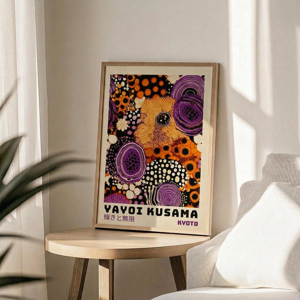 May include: Framed colorful abstract art print with purple circles, orange dots, and black polka dot patterns displayed on a light wood round side table next to a beige upholstered chair. Natural light streams across white walls creating soft shadows, with a green plant leaf visible in the corner.