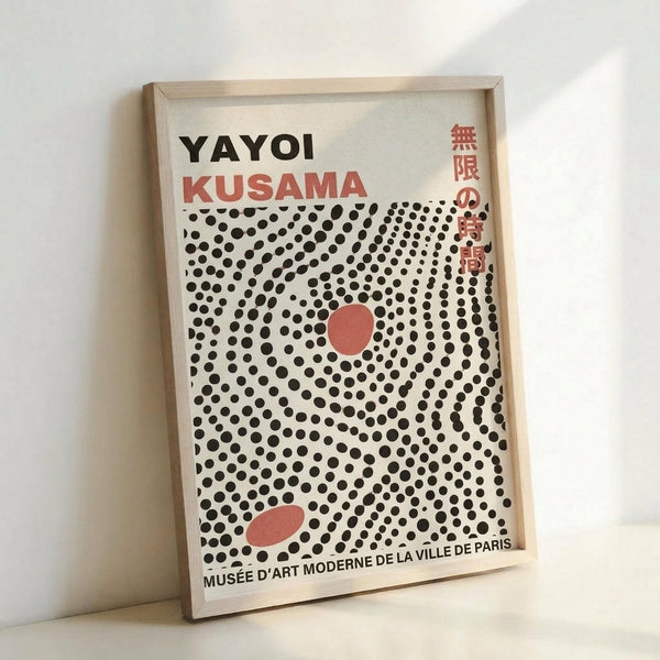 Framed poster featuring Yayoi Kusama's name in black and red text above a field of black polka dots on cream background. Two coral-red oval shapes appear among the dots. Japanese characters visible on right side. Text at bottom reads "Musée d'Art Moderne de la Ville de Paris." Light wooden frame displayed against white wall, showing dimensional depth and shadow.