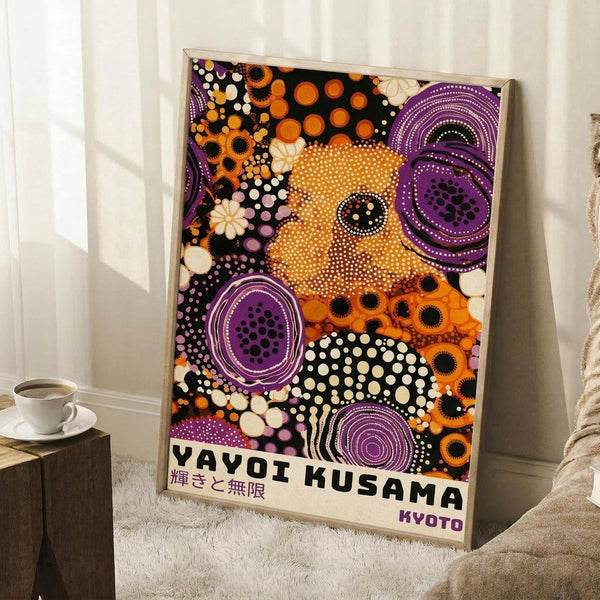 May include: Framed poster featuring vibrant dotted artwork in purple, orange, black and white with circular patterns and polka dots in Kusama's signature style. Text reads "Yayoi Kusama" with Japanese characters and "Kyoto" below. Frame leans against white wall next to coffee cup on wooden surface and beige fabric, showing scale and home decor context.