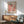 May include: Framed Yayoi Kusama exhibition poster featuring red, orange and white polka dots on cream background, displayed above wooden sideboard with white ceramic lamp. Mid-century modern living room setting with sage green upholstered armchair, round coffee table, and natural sunlight casting geometric shadows on white wall.