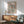 May include: Framed Yayoi Kusama exhibition poster with black polka dot pattern and coral accents on cream background, displayed above mid-century wooden sideboard with ceramic lamp. Sage green upholstered armchair with wooden frame sits beside small round coffee table. Natural sunlight creates geometric shadows on white wall in modern living room setting.