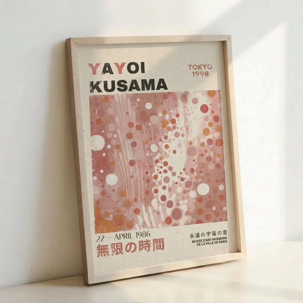 May include: Framed museum exhibition poster featuring Yayoi Kusama's signature polka dot pattern in soft pink, coral, and white tones with flowing vertical lines. The poster displays "YAYOI KUSAMA" in bold letters, "TOKYO 1998" in the corner, and Japanese text at the bottom with "22 APRIL 1986." The light wood frame leans against a white wall on a white surface, showing the poster's full layout and muted, artistic color palette reminiscent of Kusama's infinity room