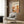 May include: Framed vintage-style art poster with orange and cream botanical design featuring abstract flowers and Japanese text, displayed on wood paneled wall above beige upholstered armchair with geometric patterned pillow, beside brass floor lamp and dark round side table with open book