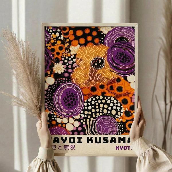 May include: Person in cream sweater holding framed poster with colorful abstract dot art in orange, purple, black and white circular patterns. Japanese text visible at bottom. Dried pampas grass and neutral curtains in background. Light wooden frame around vibrant contemporary artwork with polka dots and concentric circles in psychedelic style.