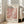 May include: Framed art poster with abstract pink and coral polka dot design, featuring "YAYOI KUSAMA" text and Japanese characters, leaning against white wall next to beige upholstered sofa in bright living room with large windows and wooden floors
