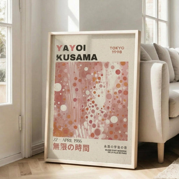 May include: Framed art poster with abstract pink and coral polka dot design, featuring "YAYOI KUSAMA" text and Japanese characters, leaning against white wall next to beige upholstered sofa in bright living room with large windows and wooden floors