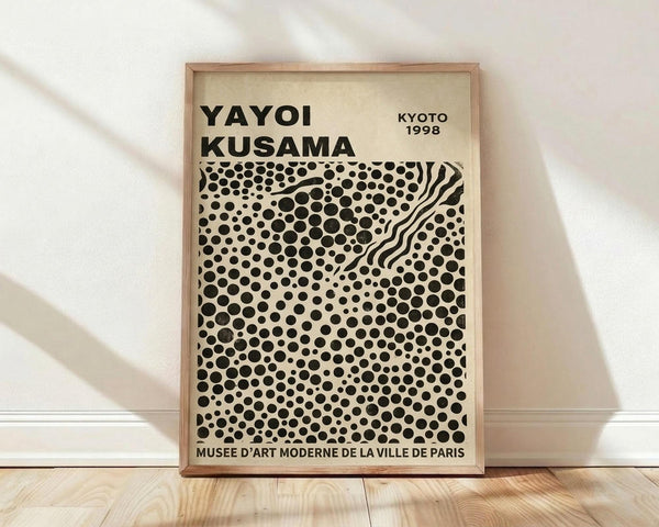 May include: Framed museum exhibition poster leaning against white wall on light wood floor. Features bold black polka dot pattern on cream background with wavy linear elements cutting through the dots. Black text at top and bottom, wooden frame, soft natural lighting creating diagonal shadows across the scene.