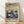 May include: Framed poster print leaning against white wall on wooden floor. Artwork features dreamy botanical scene with cream-colored mushrooms and dandelion seed heads against blue and beige swirling sky dotted with white spots. Japanese text appears at bottom. Natural wood frame with light wood grain visible. Soft natural lighting creates gentle shadows on textured white wall.