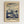 May include: Framed art print hanging on white wall featuring stylized botanical illustration with cream and blue tones. Shows abstract mushrooms, dandelions, and plants beneath swirling sky with white dots resembling stars or snow. Text reads "YAYOI KUSAMA" and "KYOTO" with Japanese characters below. Natural wood frame with cream matting creates clean, modern gallery wall appearance.