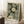 May include: Framed art print leaning against white wall showing green background with white polka dot patterns forming dandelion-like circular flowers. Text reads "YAYOI KUSAMA" and Japanese characters with "KYOTO" below. Light wood frame, displayed on white fluffy rug next to wooden block with coffee cup, cream-colored throw blanket visible, natural window light creating shadows on wall.
