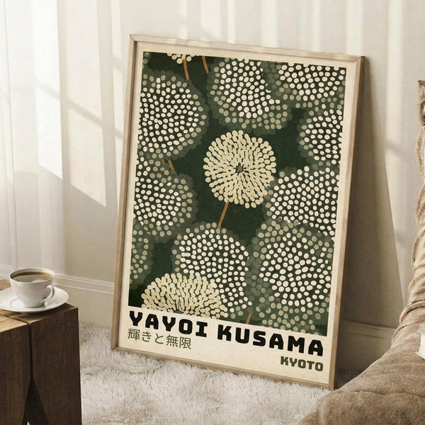 May include: Framed art print leaning against white wall showing green background with white polka dot patterns forming dandelion-like circular flowers. Text reads "YAYOI KUSAMA" and Japanese characters with "KYOTO" below. Light wood frame, displayed on white fluffy rug next to wooden block with coffee cup, cream-colored throw blanket visible, natural window light creating shadows on wall.