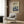 May include: Framed poster with dreamy blue and cream artwork featuring mushroom-like forms under swirling sky, displayed above modern beige armchair with geometric pillow in contemporary room setting with wood paneling, brass floor lamp, and dark round side table with open book.