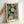 May include: A framed art print featuring dotted dandelion-like flowers in cream and white against a dark green background, displayed in a light wood frame. A hand reaches toward the left side of the frame for scale reference. The artwork shows organic circular patterns made of small dots, with "YAYOI KUSAMA" and "KYOTO" text at the bottom along with Japanese characters. The print rests against a white wall.