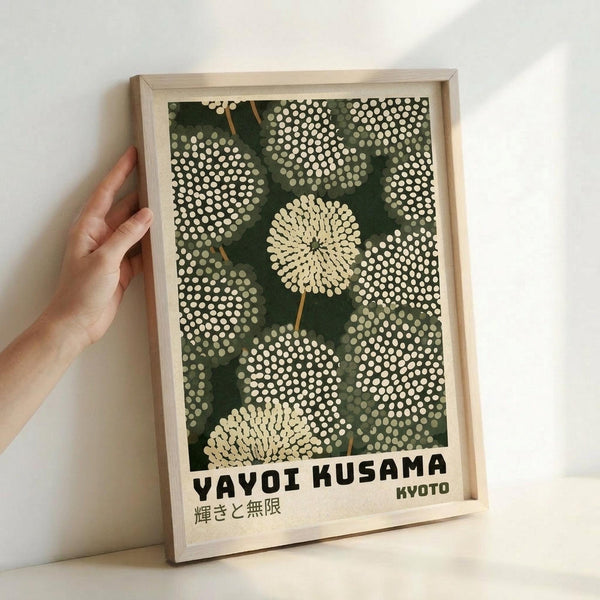 May include: A framed art print featuring dotted dandelion-like flowers in cream and white against a dark green background, displayed in a light wood frame. A hand reaches toward the left side of the frame for scale reference. The artwork shows organic circular patterns made of small dots, with "YAYOI KUSAMA" and "KYOTO" text at the bottom along with Japanese characters. The print rests against a white wall.