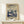 May include: Framed art print displayed on light wood shelf against cream wall, featuring stylized mushrooms and flora in blue and cream tones with swirling sky dotted with white spots, Japanese text at bottom, small wrapped white gift box with greenery beside frame