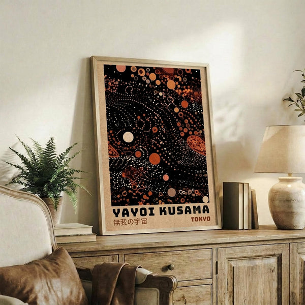 May include: Framed poster with black background featuring scattered circles in orange, coral, and cream tones inspired by Yayoi Kusama's dot art, displayed on rustic wooden sideboard next to fern plant, ceramic vase, and books in warm-toned living room setting