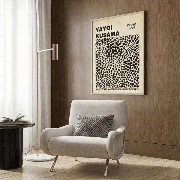 May include: A cream-colored poster with black polka dot pattern and wavy lines mounted on wood-paneled wall above beige upholstered armchair with geometric patterned pillow. Modern floor lamp with brass finish and white shade stands nearby next to dark round side table with open book. Natural light filters through sheer curtains, creating warm ambient lighting in contemporary living room setting.