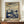 May include: Framed art print displayed on wooden sideboard next to white ceramic vase and stacked books. The poster features cream and blue tones with flowing wavy sky, white dots throughout, and silhouettes of mushrooms and botanical elements in foreground. Text reads "YAYOI KUSAMA" and "KYOTO" at bottom with Japanese characters. Soft natural lighting in elegant room setting with white walls and crown molding.