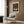 May include: Framed art poster featuring a stylized tree with colorful dotted foliage against a gradient sunset background, displayed on wood paneling above a beige upholstered armchair with geometric patterned pillow, alongside a brass floor lamp and dark side table with open book.
