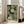 May include: Framed poster with green background featuring cream-colored polka dot pattern resembling dandelions or pom-poms, with Japanese text and "YAYOI KUSAMA" and "KYOTO" in black letters at bottom. Light wood frame leaning against white wall next to beige upholstered sofa in bright living room with large windows.