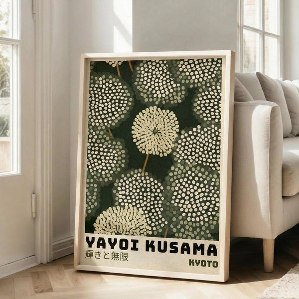 May include: Framed poster with green background featuring cream-colored polka dot pattern resembling dandelions or pom-poms, with Japanese text and "YAYOI KUSAMA" and "KYOTO" in black letters at bottom. Light wood frame leaning against white wall next to beige upholstered sofa in bright living room with large windows.