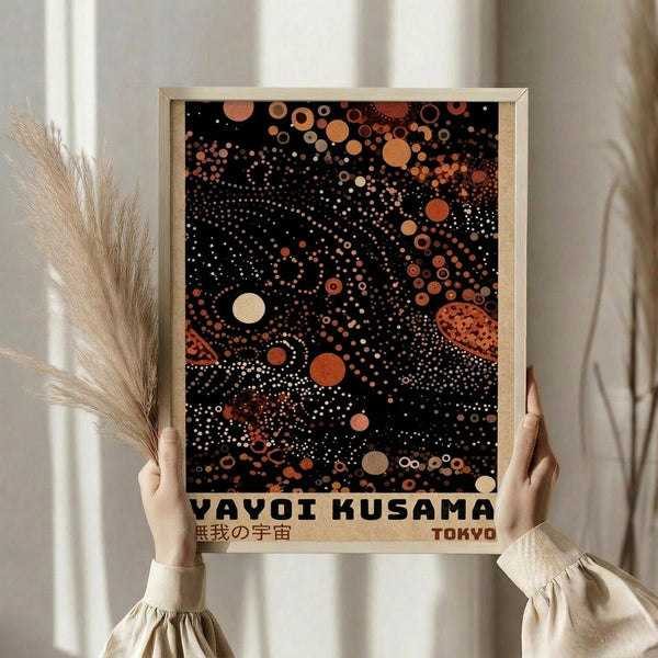 May include: Person in cream sweater holding framed art print featuring black background with orange, coral, and white polka dots in cosmic pattern. Japanese text below reads "YAYOI KUSAMA" and "TOKYO" with characters. Light wood frame, dried pampas grass visible, soft natural lighting against white curtained window background.