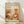 May include: Framed poster with cream background featuring "YAYOI KUSAMA" text and Japanese characters, displaying abstract botanical illustration in warm orange, brown, and cream tones with stylized flowers, leaves, and dotted patterns. Wood frame leaning against white wall on light wooden floor with natural sunlight casting shadows.