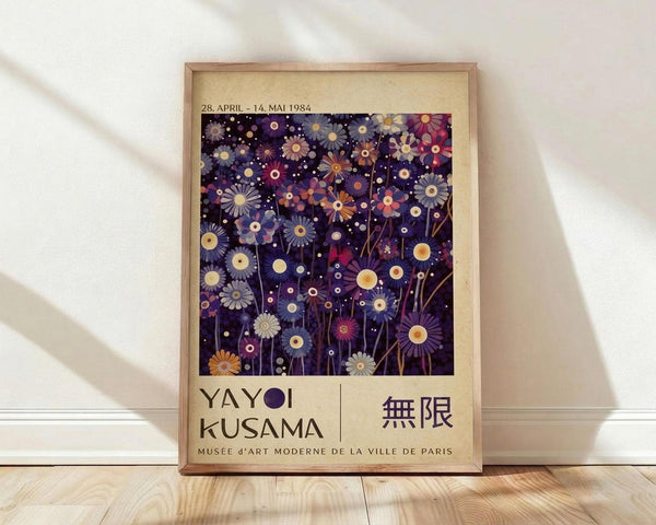 May include: A framed vintage exhibition poster featuring stylized purple, blue, and orange daisy-like flowers scattered across a dark background with white dots. The cream-colored poster shows "YAYOI KUSAMA" and Japanese characters below the floral artwork, along with museum exhibition dates from 1984. The wooden frame leans against white walls on light wooden flooring with natural sunlight casting diagonal shadows.