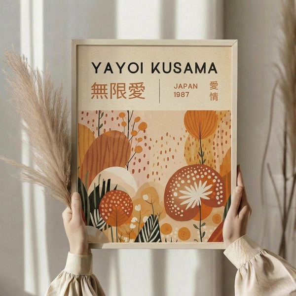 May include: Person holding framed vintage-style poster with botanical illustration in warm oranges, browns, and creams. Features stylized flowers, leaves, and geometric patterns. Japanese text visible at top. Light wooden frame against neutral curtained background. Dried pampas grass stems visible on left side.
