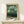 May include: Framed art print on light wood shelf against cream wall, featuring stylized trees and botanical elements in green tones with dotted patterns and circular shapes. Small white wrapped gift box with twine sits beside the natural wood frame. The artwork shows an abstract forest scene with whimsical polka-dotted foliage in sage green and cream colors.