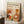 May include: Framed poster with "YAYOI KUSAMA" text at top, Japanese characters, and "JAPAN 1987" below. Abstract illustration features warm orange, yellow, and coral circular shapes, botanical elements like flowers and leaves, and polka dot patterns in mid-century modern style. Wooden frame leans against white wall corner near cozy textured blanket and coffee cup on wooden surface.