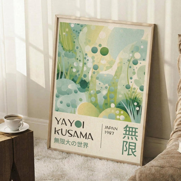May include: Framed art print with abstract green and white botanical design featuring organic shapes, circles, and flowing forms in watercolor style. Shows "YAYOI KUSAMA" and "JAPAN 1987" text with Japanese characters at bottom. Light wood frame leaning against white wall near window, with coffee cup on wooden surface and textured throw blanket visible in cozy interior setting.