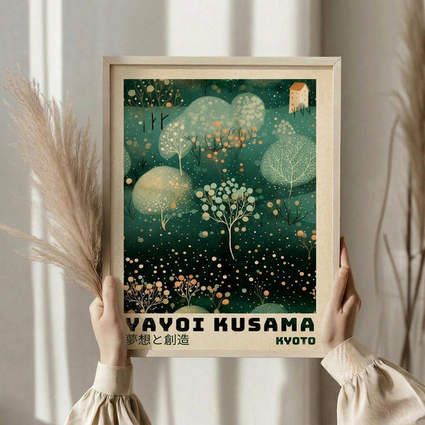 May include: Person in cream sweater holding framed art print in light wooden frame. Green botanical artwork features abstract trees, circular shapes, and dots in various shades of green with cream accents. Text reads "YAYOI KUSAMA" and "KYOTO" in black letters at bottom. Dried pampas grass visible on left side. Neutral background with soft natural lighting.