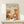 May include: Framed art print displaying "YAYOI KUSAMA" text at top with Japanese characters and "JAPAN 1987" below. Abstract botanical illustration features warm orange, rust, and cream colored organic shapes, polka-dotted mushrooms, stylized plants and flowers in mid-century modern style. Natural wood frame sits on light wooden surface against cream wall with small wrapped gift box nearby.